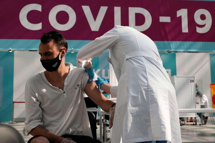 Медицинский работник в белом халате и перчатках делает прививку сидящему мужчине в маске.