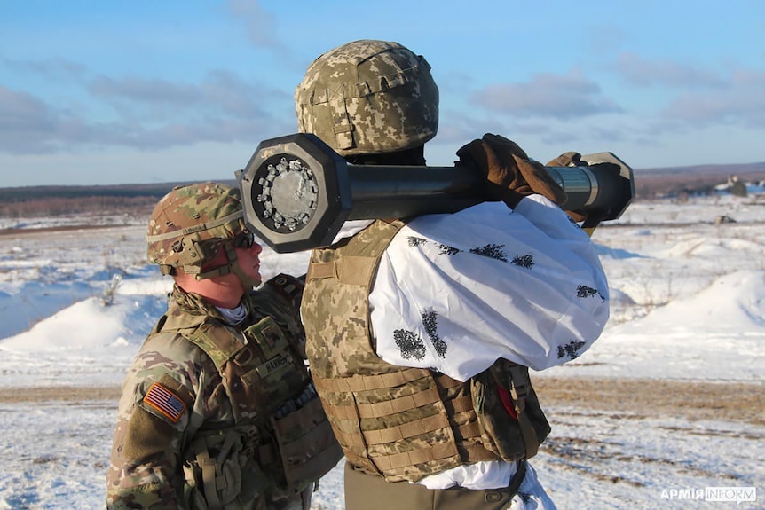 Американский инструктор в военной экипировке стоит позади украинского солдата, держащего в руках ракету M141 Bunker Defeat Munition.