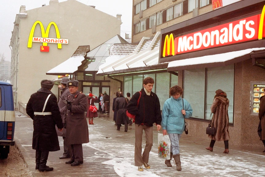 Люди проходят мимо охранников в советской форме перед рестораном быстрого питания с красными и желтыми брендами.